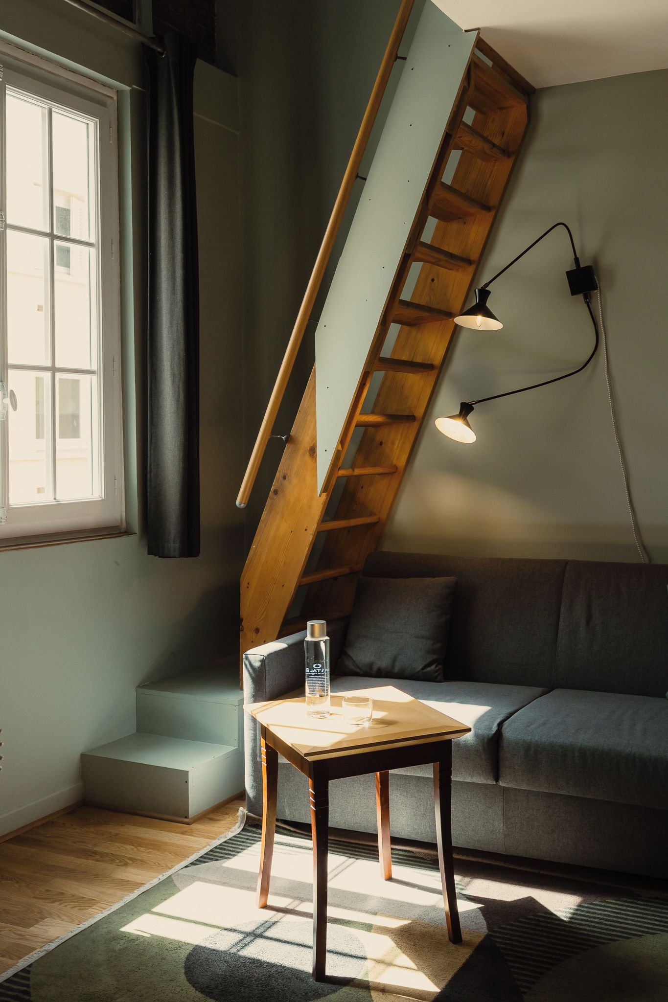 Een loft-achtige kamer met een schuine houten trap, een bank, een tafel met fles en glas, en een wandlamp.