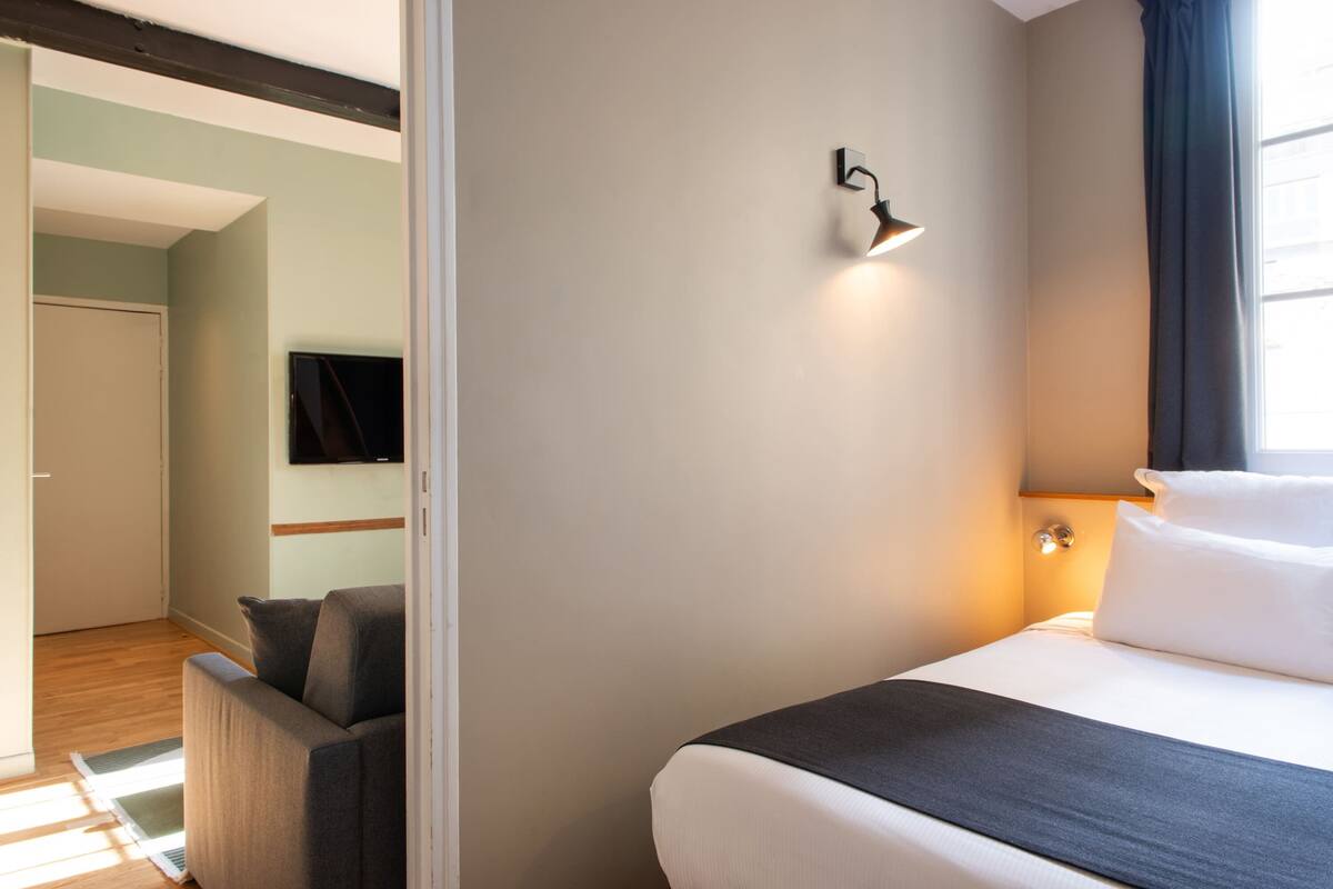 A hotel room with a bed, a sofa, a flat-screen TV mounted on the wall, and a window with curtains.