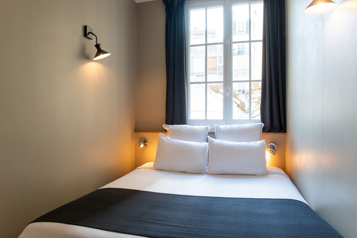 A neatly made bed with white linens and a dark throw blanket, a window with dark curtains, and a wall-mounted light fixture.