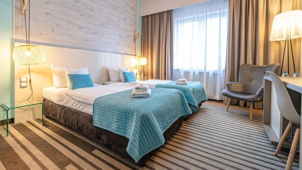 A hotel room with two beds, a desk, a chair, and a large window with curtains. - NoBo Hotel (Lodz)