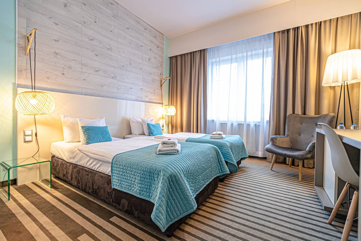 Habitación de hotel con dos camas, un escritorio, una silla y un amplio ventanal con cortinas.