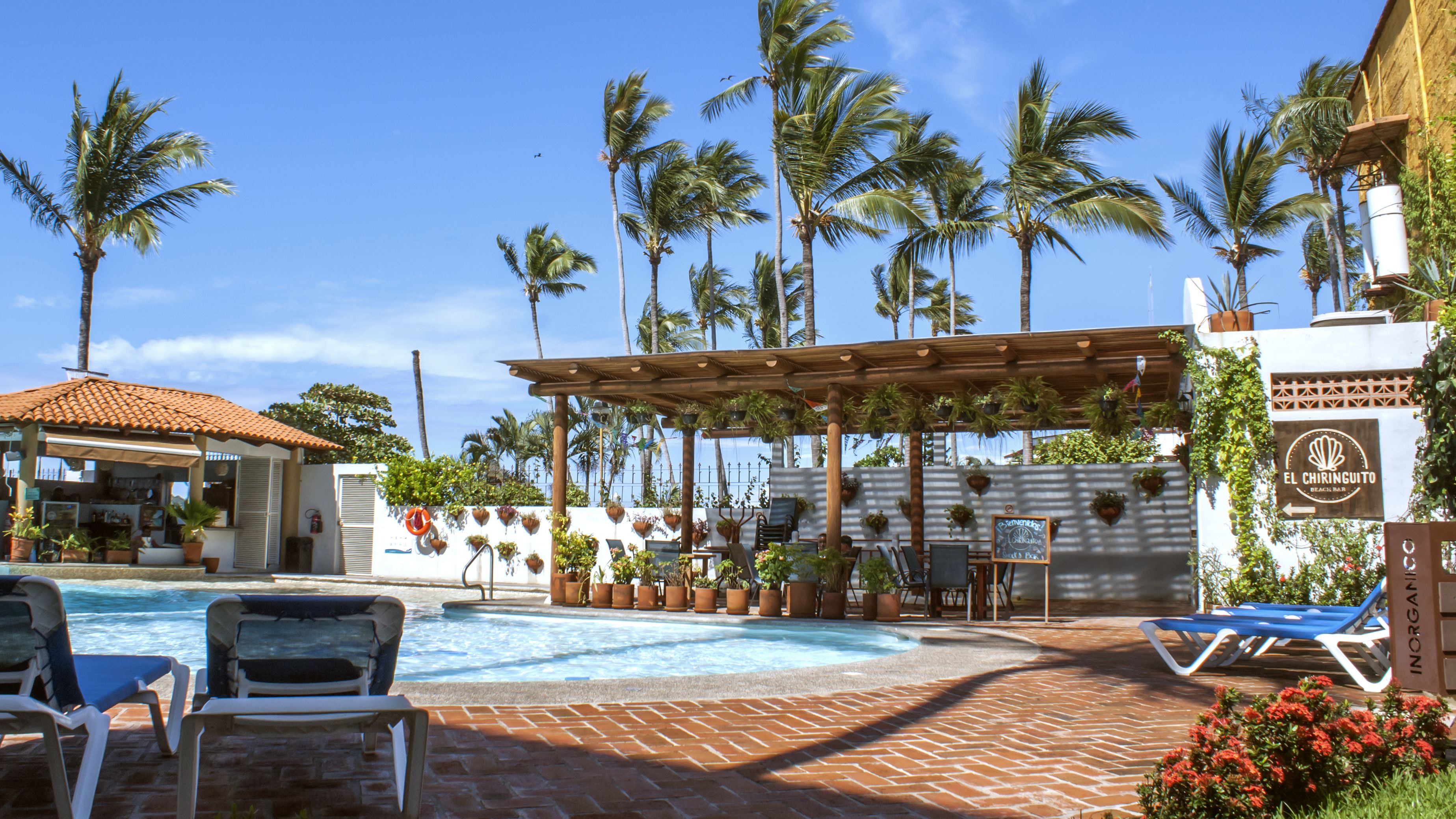 Un espace piscine avec des chaises longues, une pergola ornée de plantes suspendues et un panneau indiquant « El Chiriguito Beach Club ».