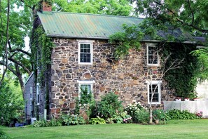 A stone house with two windows and a metal roof, surrounded by lush greenery and a white fence.