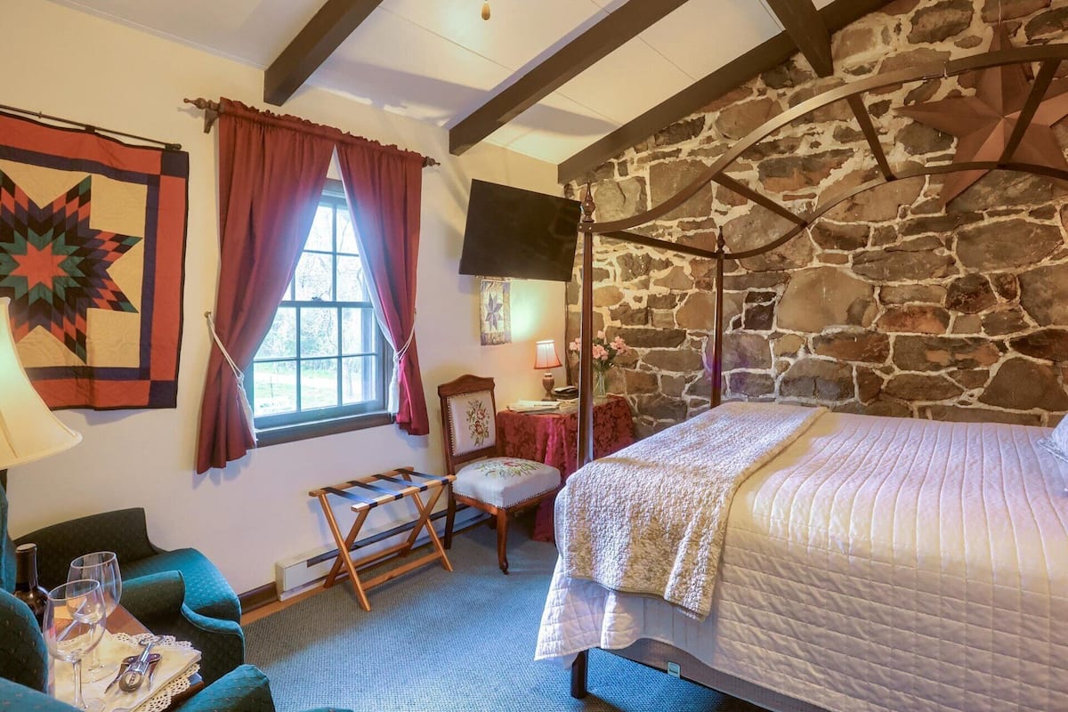 A cozy room with a stone wall, a four-poster bed, a window with curtains, a chair, and a small table with wine glasses.