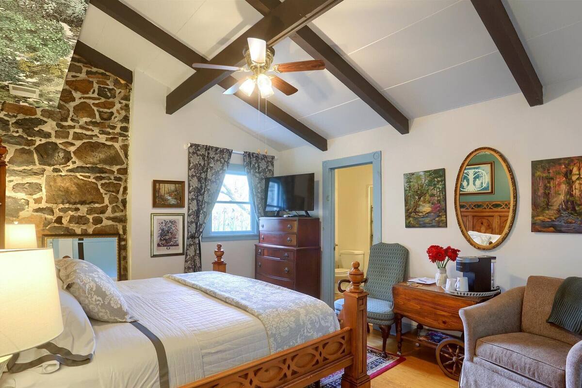A bedroom with a stone wall, a bed with a wooden headboard, a ceiling fan, a dresser, a chair, a desk with a computer, and artwork on the walls.
