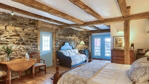 A rustic bedroom with exposed wooden beams, a stone wall, a large bed with a quilt, a wooden chair, and a desk with a vase of flowers. - Battlefield Bed & Breakfast Inn (Gettysburg)