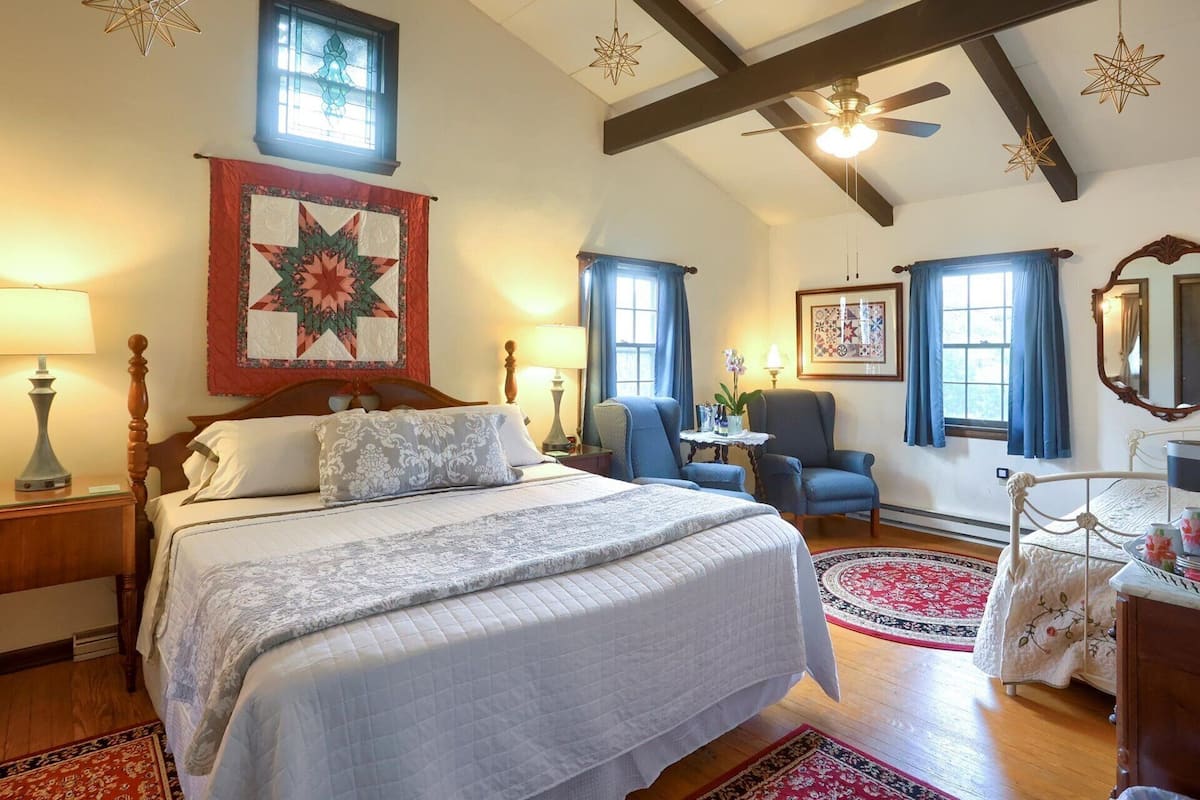A bedroom with a large bed, a ceiling fan, a window with curtains, and a quilt hanging on the wall.