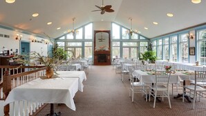 A spacious dining area with high ceilings, large windows, and a central fireplace. Tables are set with white tablecloths and chairs. - Battlefield Bed & Breakfast Inn (Gettysburg)