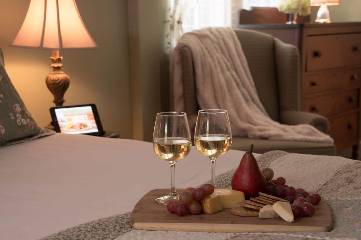 A bed with a wooden board holding glasses of white wine, a pear, grapes, and cheese.