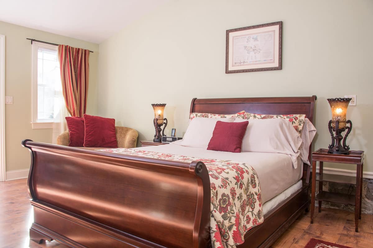 A bedroom with a wooden bed, red pillows, a floral bedspread, two bedside tables with lamps, a framed picture on the wall, and a window with curtains.