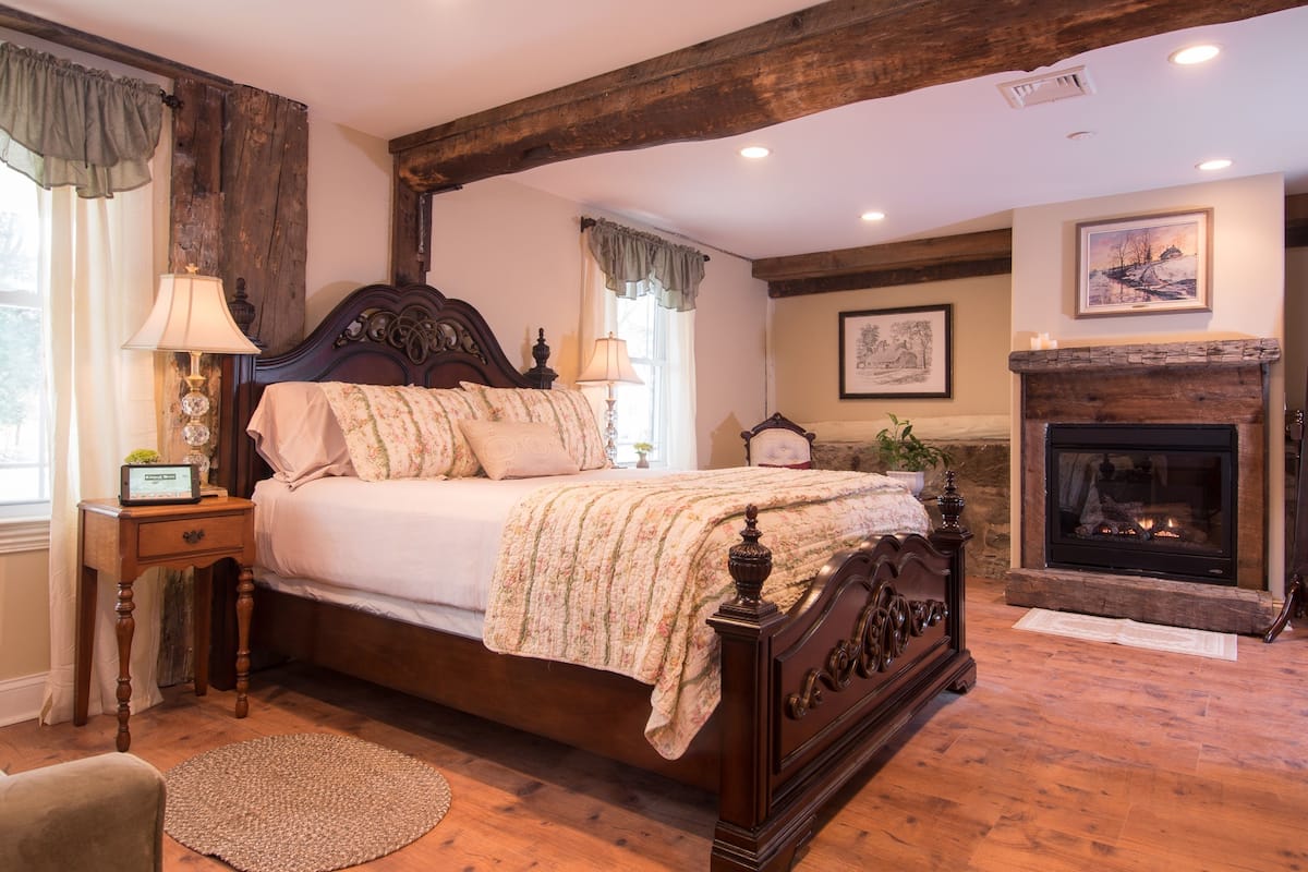 A bedroom with a large bed, wooden beams, a fireplace, and a window with curtains.