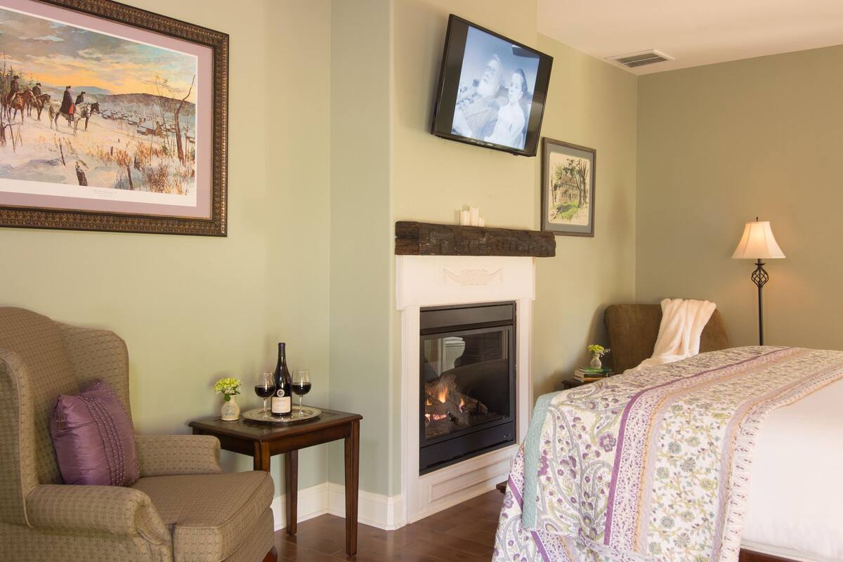 A room with a fireplace, a television mounted on the wall, a bed with a floral bedspread, and a chair with a cushion.
