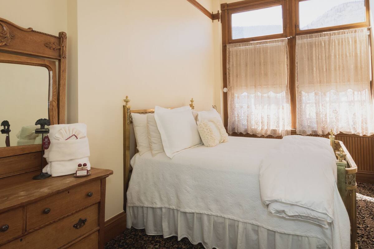 Un dormitorio con un aparador de madera, una cama con ropa blanca y una ventana con cortinas de gasa.