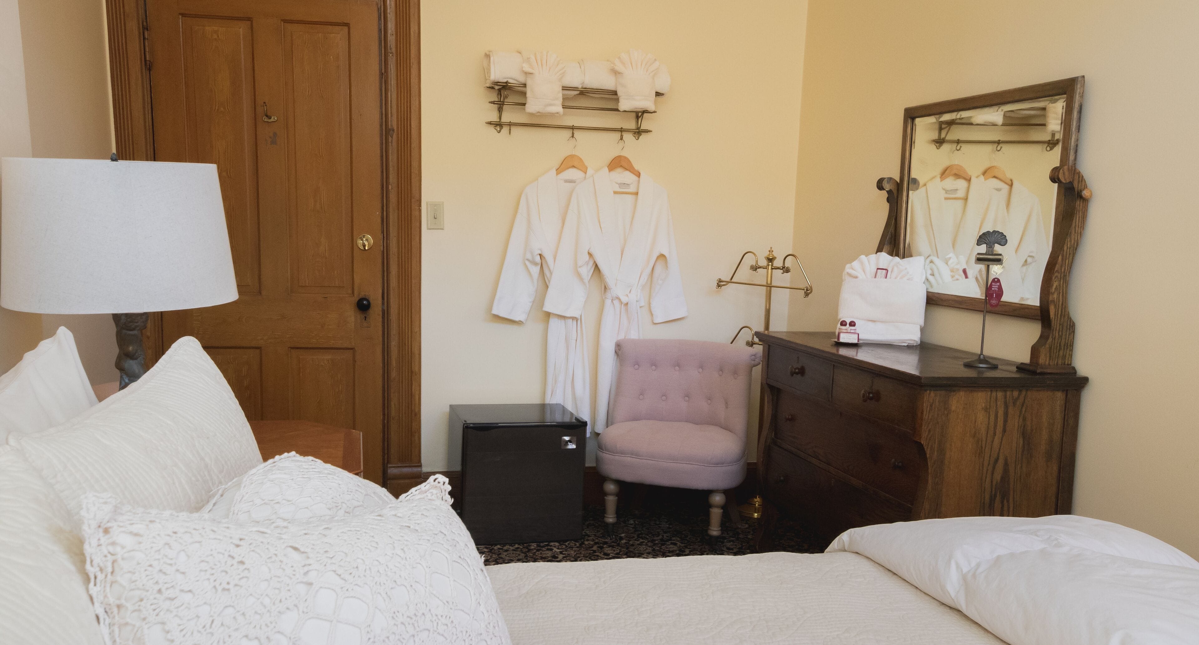 A hotel room with a bed, a dresser, a vanity with a mirror, and a chair. There are robes hanging on the wall and a lamp on the dresser.