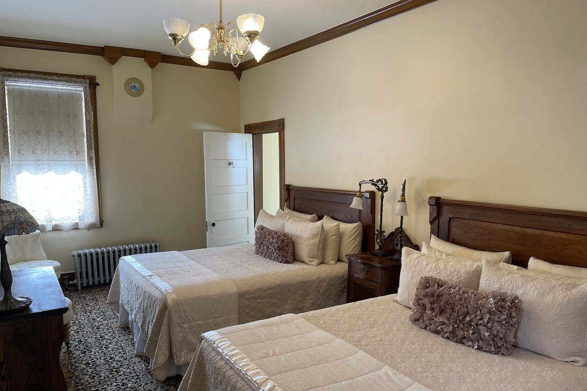 Habitación de hotel con dos camas, una mesita de noche, una lámpara, un radiador y una ventana con cortinas.