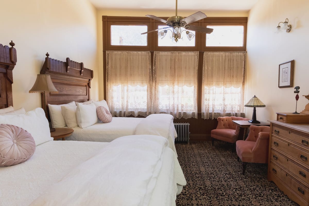 Un dormitorio con dos camas, un aparador, un escritorio, una silla, un ventilador de techo y una ventana con cortinas.