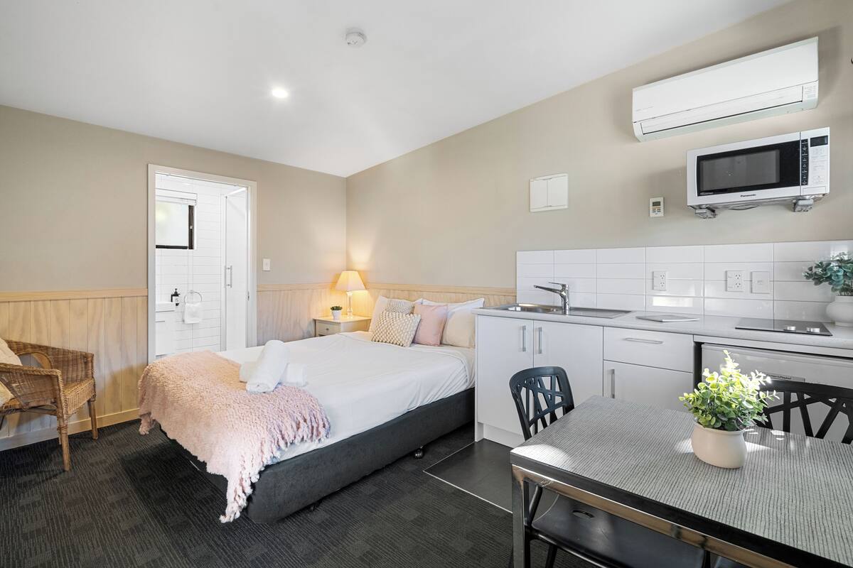 Una habitación de hotel moderna con cama, mesita de noche, lámpara y una kitchenette con microondas y fregadero.