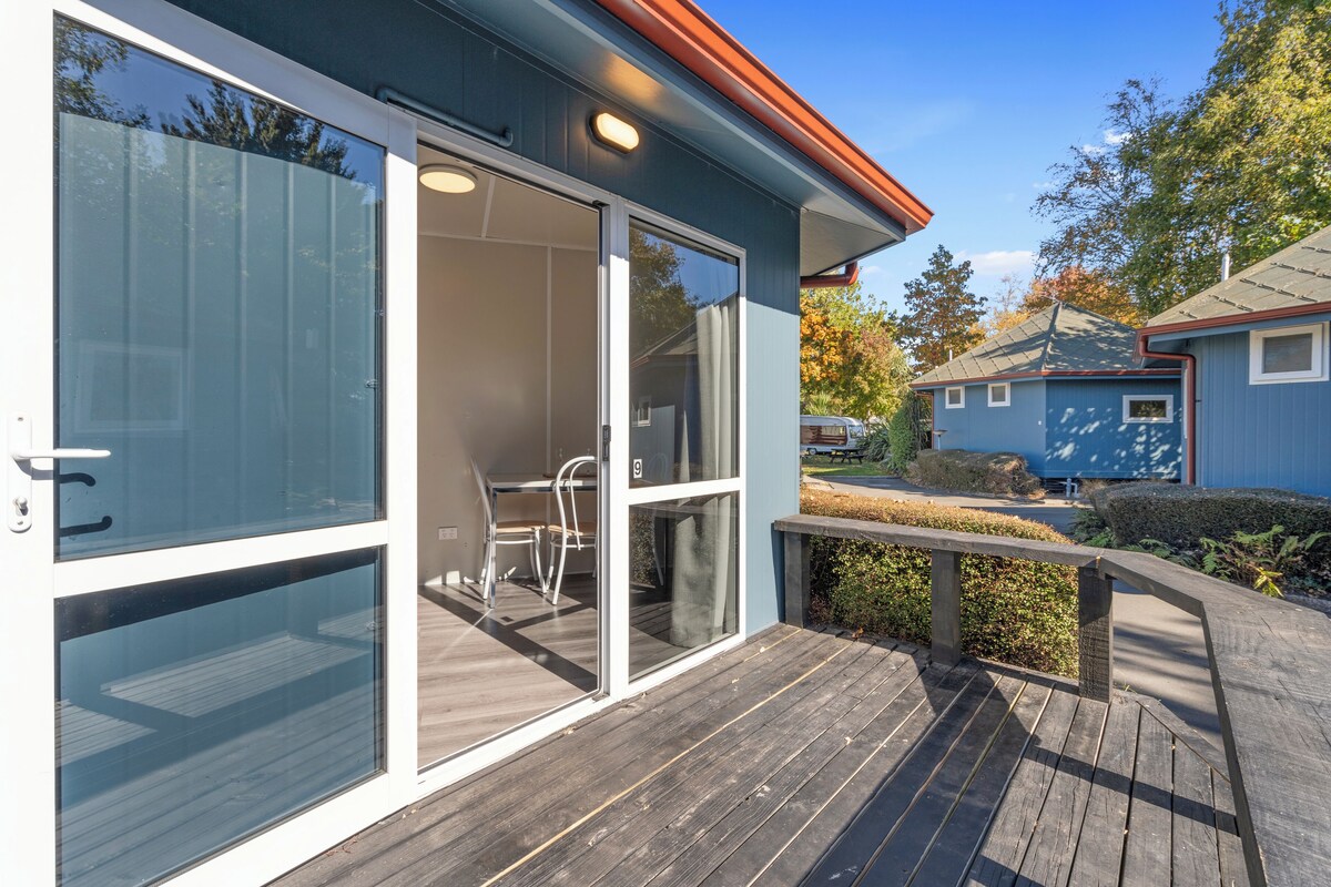 Ein modernes Haus mit einer Holzterrasse, einer Glastür, die auf eine Terrasse führt, und einer kleinen Sitzgelegenheit im Freien.