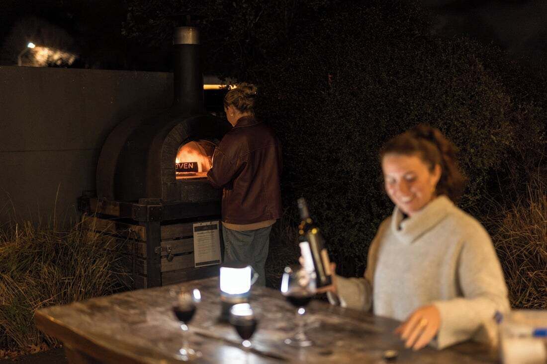 Een buitenoven voor pizza, met iemand die kookt, een tafel met wijnglazen en een fles.