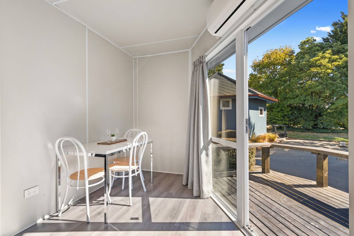 Un balcón con terraza de madera, una mesa de comedor con sillas y vistas a una casa y árboles.