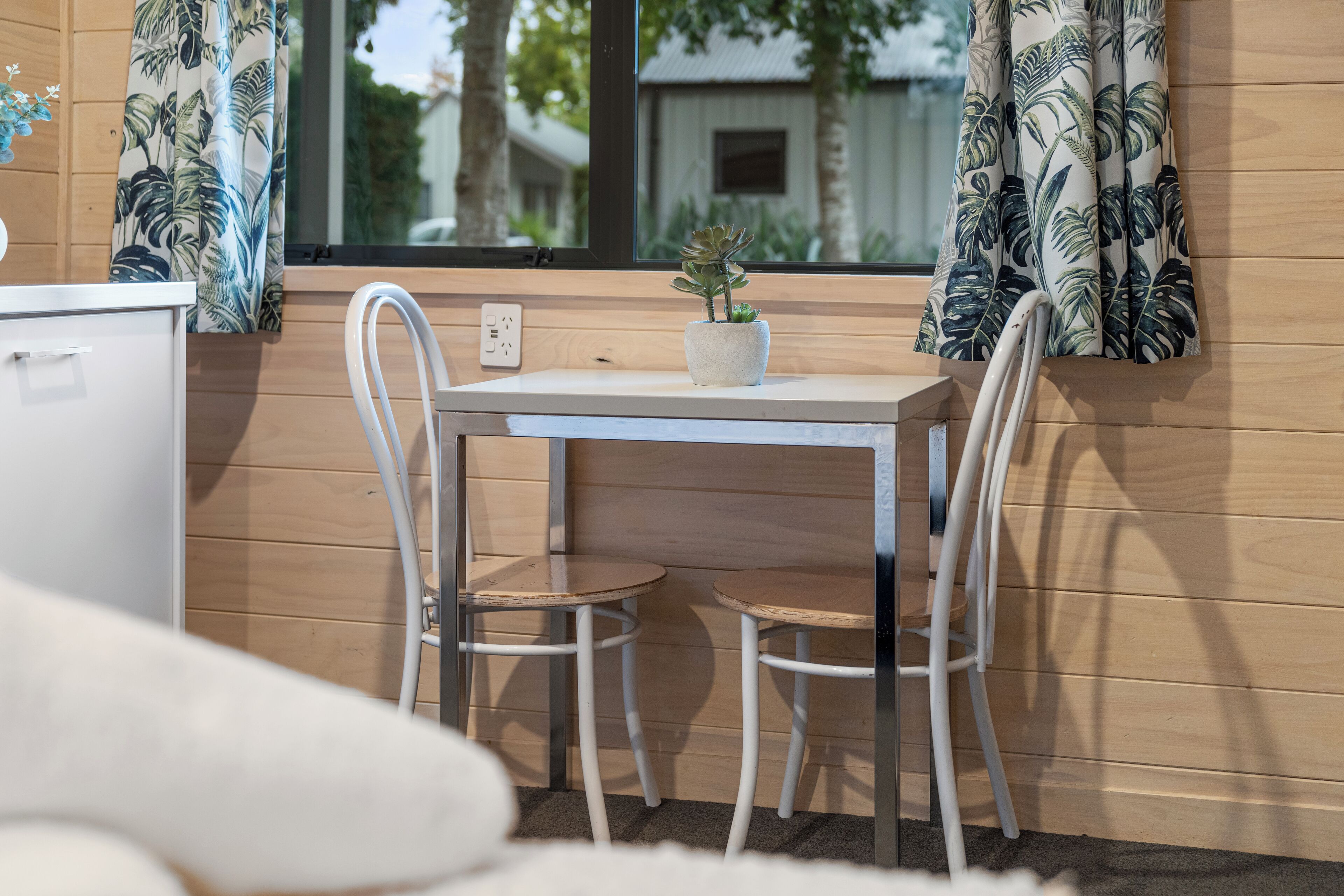 Una habitación pequeña con pared de madera, una mesa blanca con dos sillas, una planta en maceta y una ventana con cortinas.