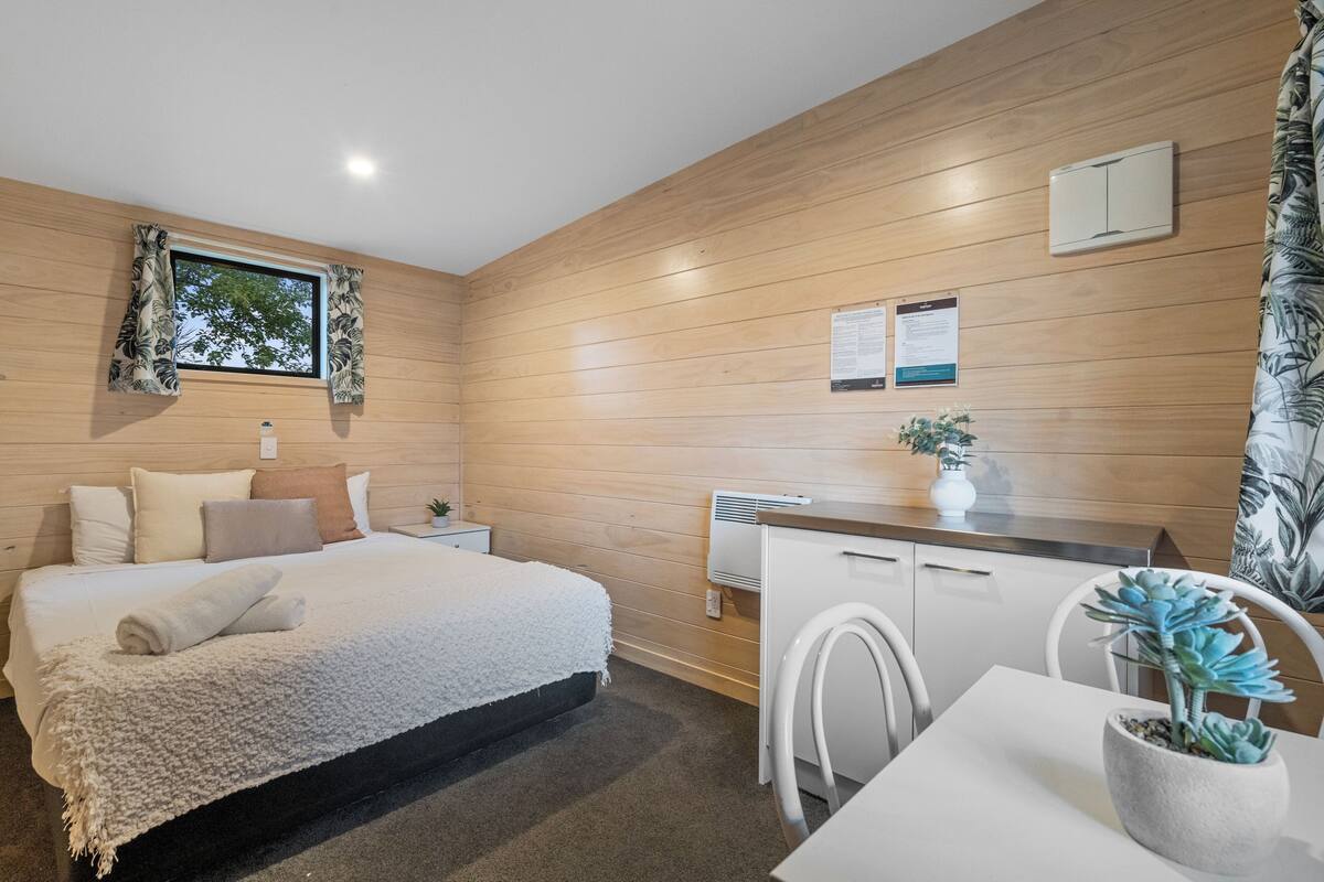 Un dormitorio moderno con una cama grande, una mesita de noche pequeña, una mesita auxiliar, una ventana con cortinas, una pared de madera, un escritorio blanco con una silla, una planta en maceta y un armario blanco.