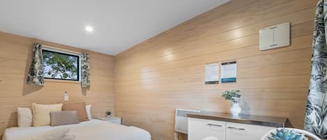 A modern bedroom with a large bed, a small bedside table, a nightstand, a window with curtains, a wooden wall, a white desk with a chair, a potted plant, and a white cabinet.