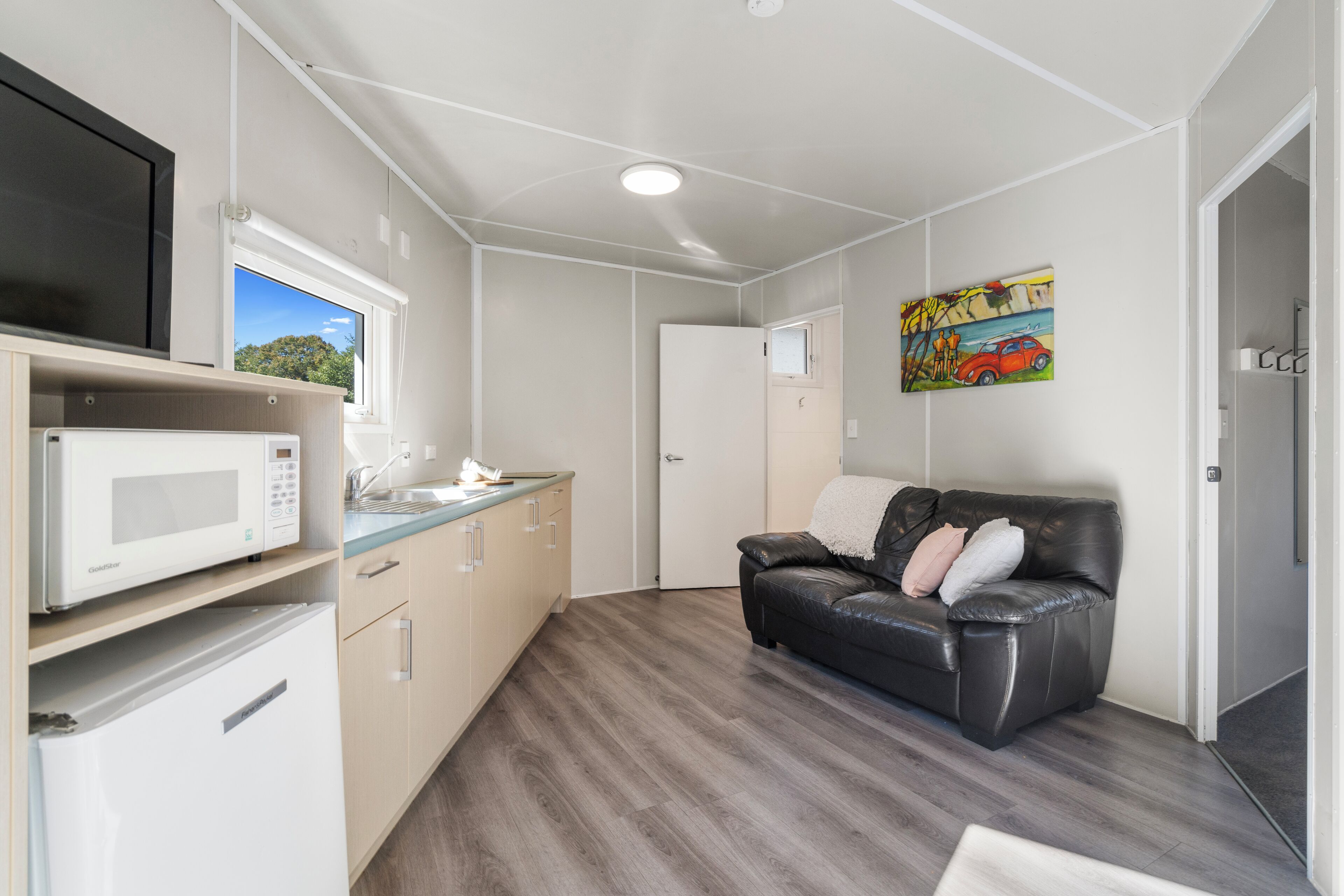 A compact kitchen with a microwave, a sink, and a window. There is a black leather sofa with cushions and a painting of a car on the wall.