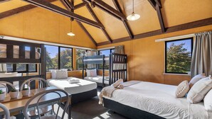 A bedroom with a large bed, a desk, and chairs. There are windows with curtains, a bunk bed, and a view of trees. - Tasman Holiday Parks - Christchurch (Christchurch)