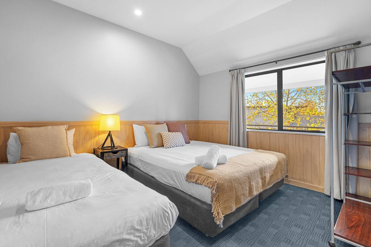 Habitación de hotel con dos camas, una mesita de noche con lámpara, ventana con cortinas y vista a árboles.