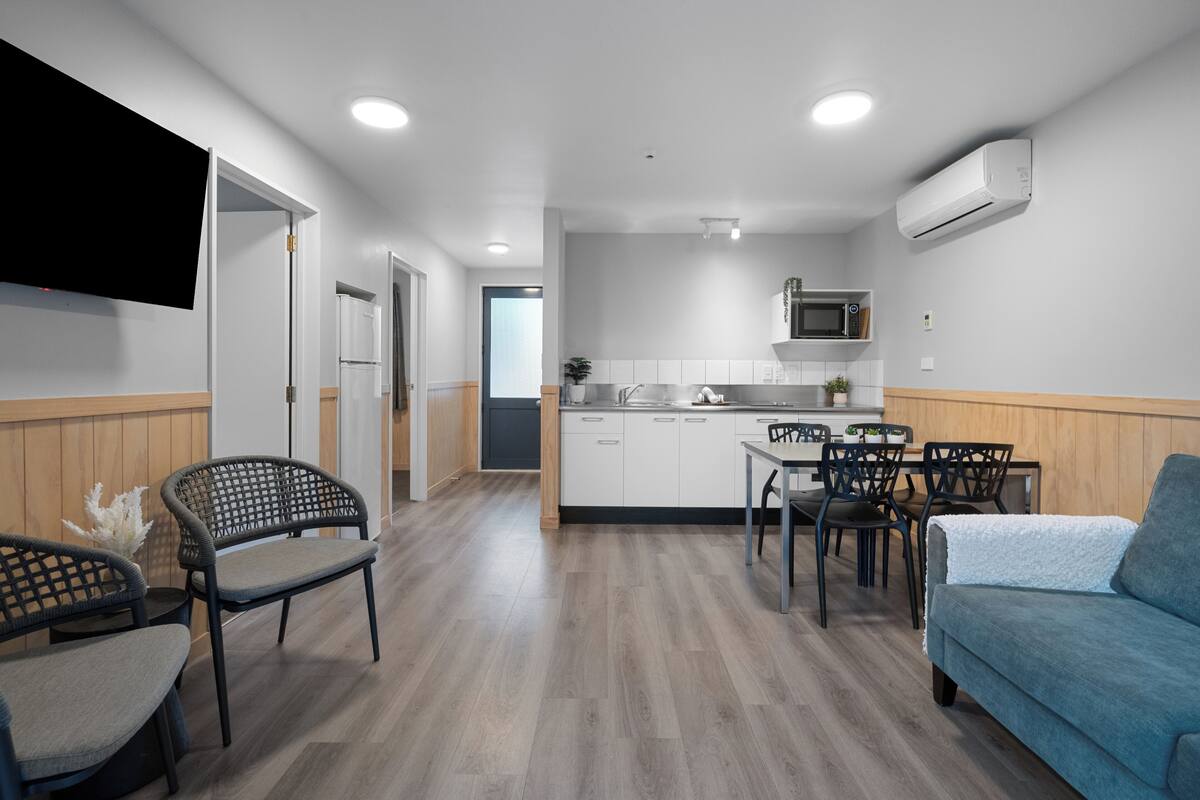 Cocina moderna con armarios blancos, microondas y equipo de aire acondicionado. Hay un comedor con mesa y sillas, y un sofá azul.
