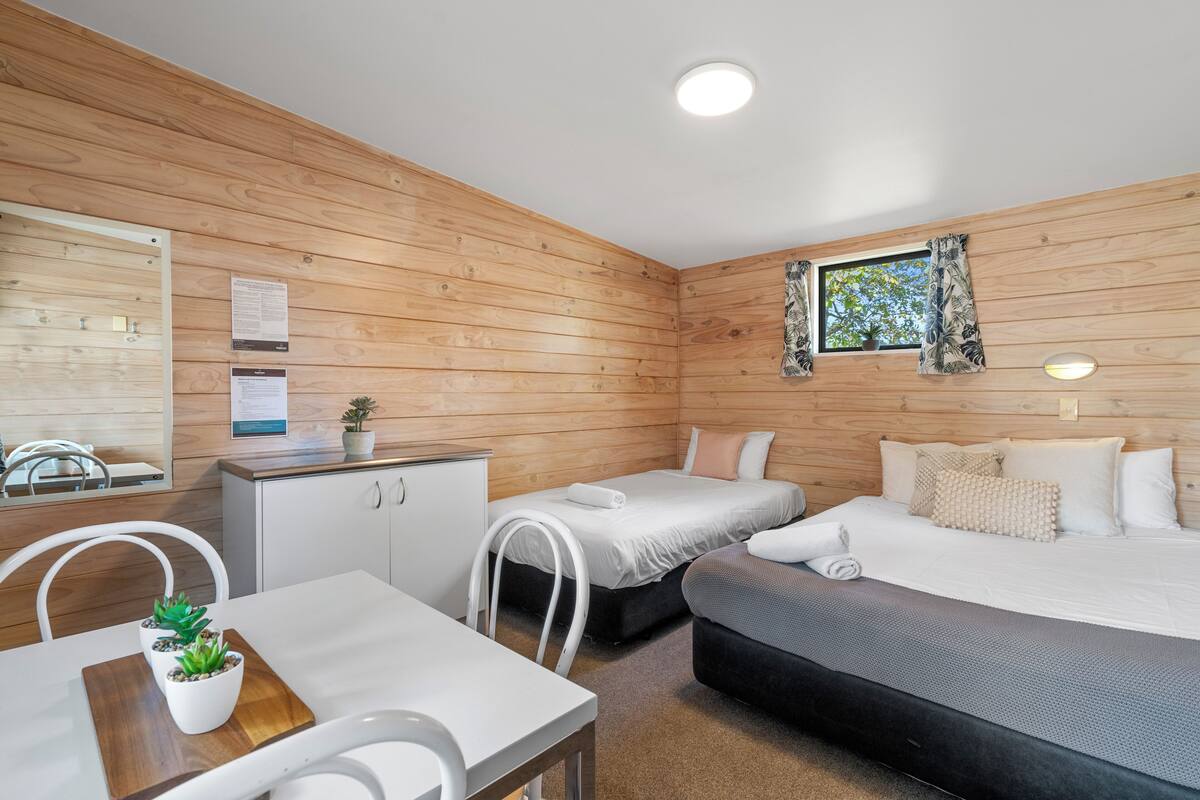 Una habitación con paredes de madera, una cama, un escritorio y una silla. Hay una ventana con cortinas y una pequeña planta en el escritorio.