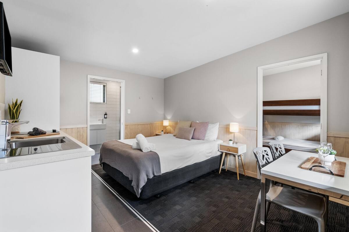 Habitación de hotel moderna con cama, mesita de noche, lámpara y una pequeña cocina con pileta y mostrador.