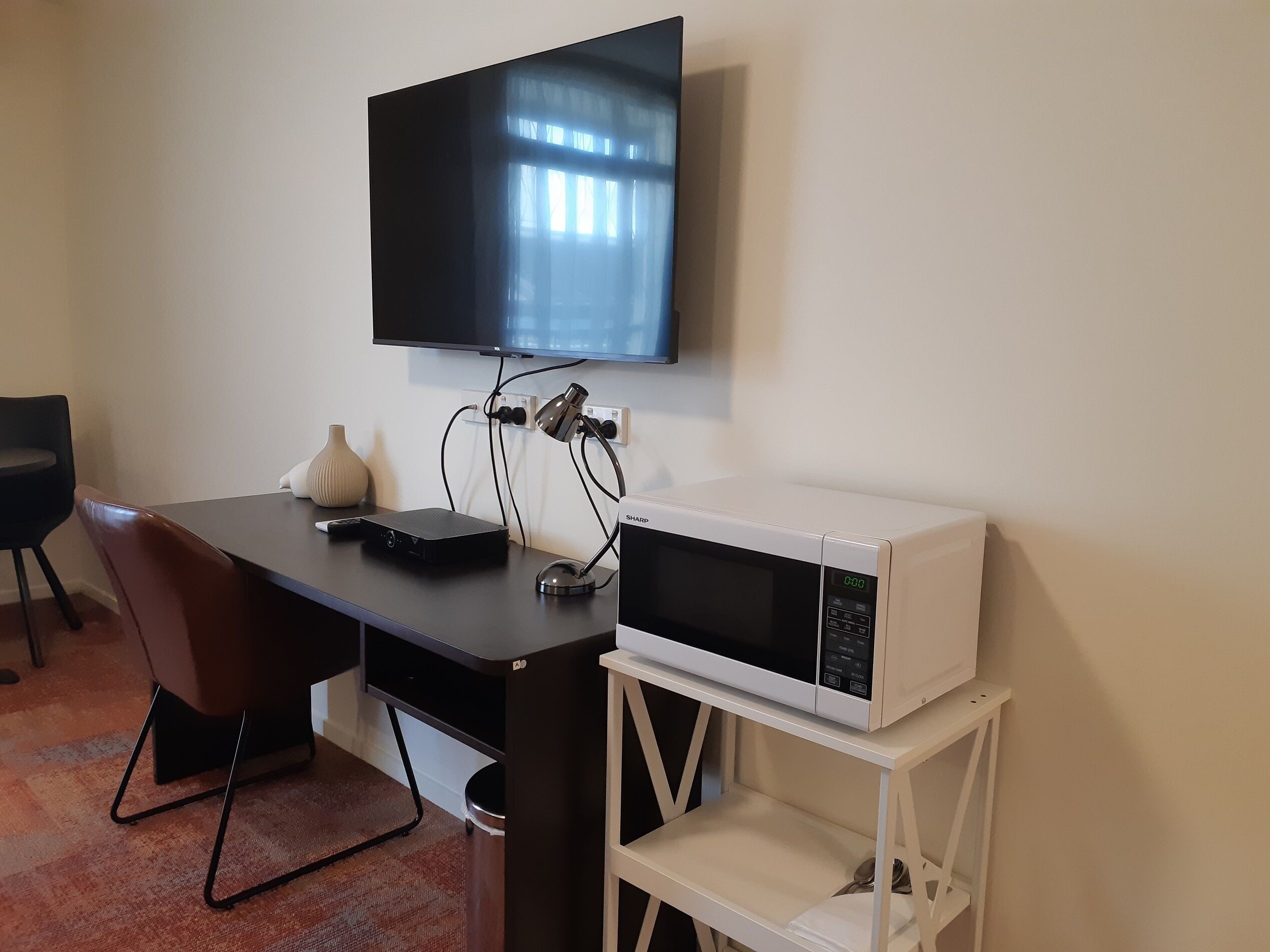 A small, modern room with a desk, a flat-screen TV mounted on the wall, a microwave on a white shelf, and a chair.