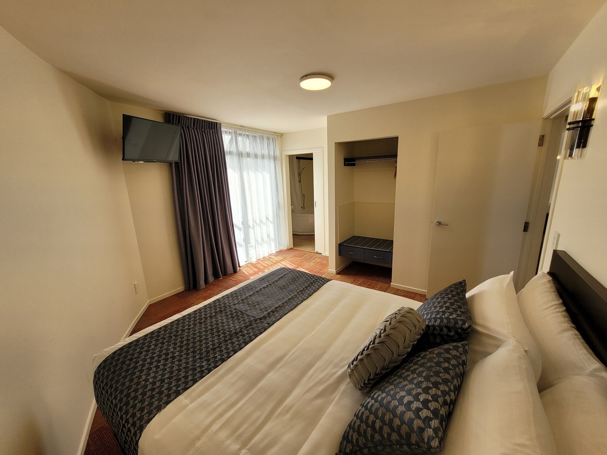 A hotel room with a bed, a TV mounted on the wall, a walk-in closet, and a window with curtains.