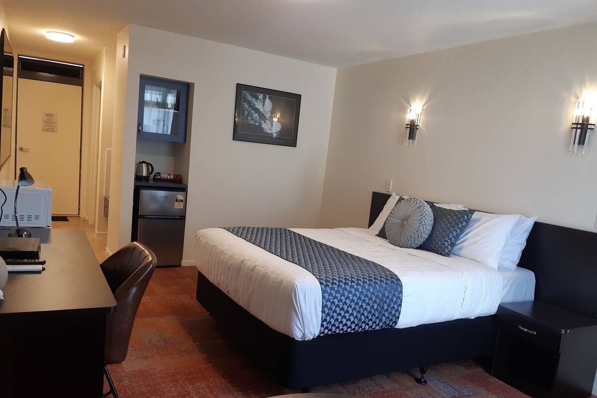 A hotel room with a bed, bedside tables, a desk with a lamp, a refrigerator, and a small kitchenette.