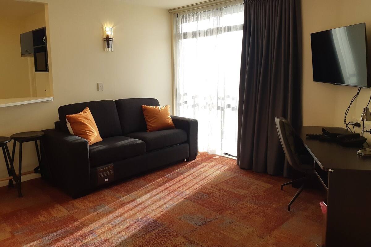 A living room with a black sofa, two orange cushions, a small table, a TV, and a large window with curtains.