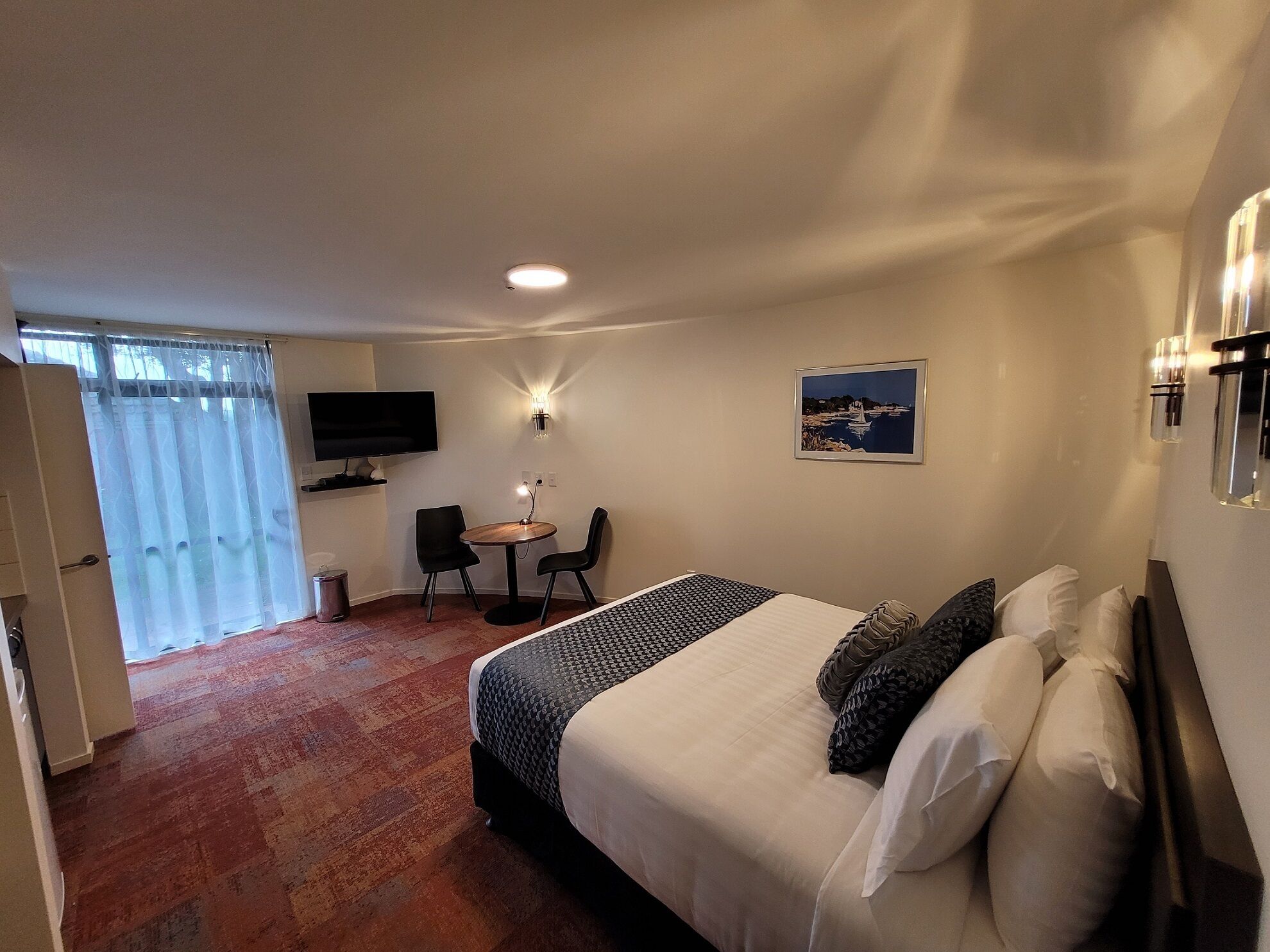 A hotel room with a bed, a desk with two chairs, a television, and a painting on the wall.
