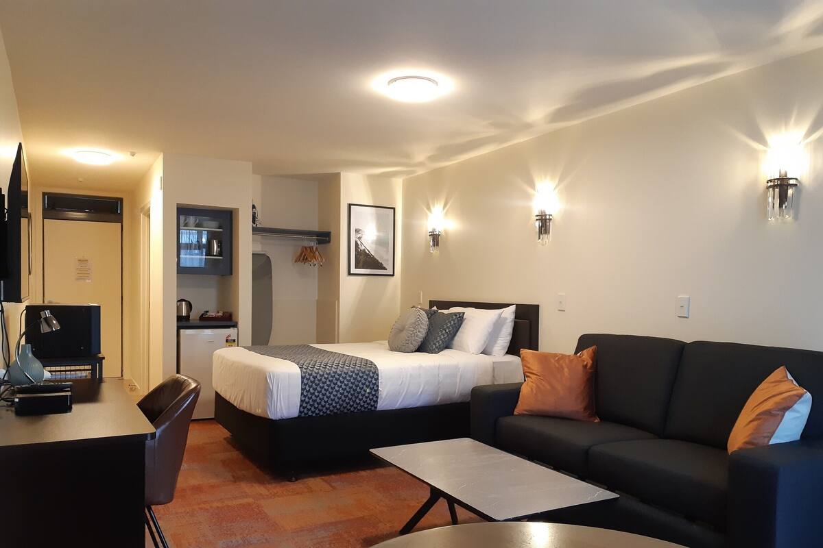 A compact hotel room with a bed, a sofa, a desk, and a kitchenette.