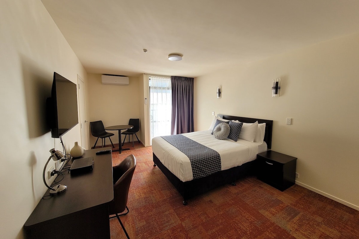A hotel room with a bed, bedside tables, a desk with a chair, a television, and a window with curtains.