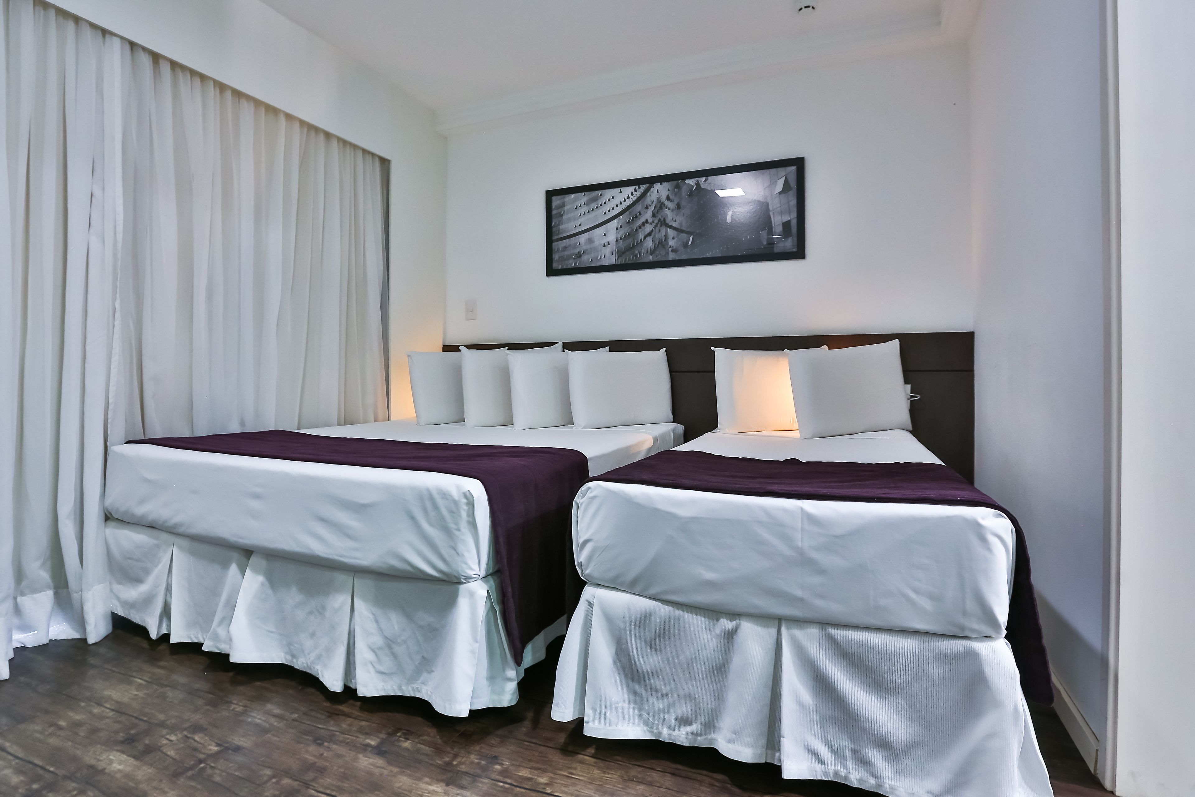 A hotel room with two beds, white bedding, a dark headboard, a framed picture on the wall, and a large window with curtains.