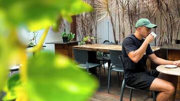 Una persona sentada en una mesa al aire libre con sillas, piso de madera y plantas en macetas al fondo.
