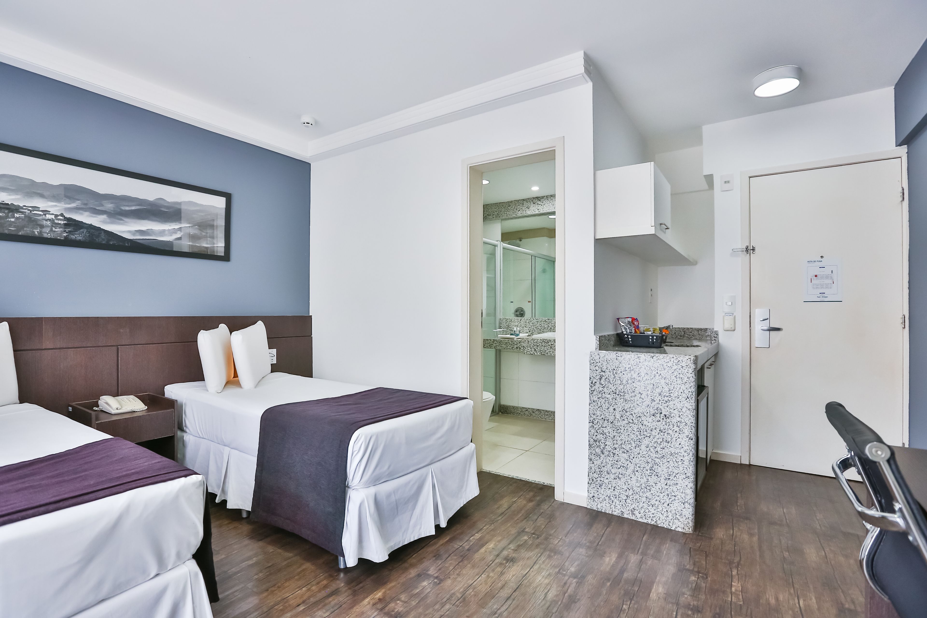Ein Hotelzimmer mit zwei Betten, einer kleinen Küchenzeile und einem durch eine offene Tür einsehbaren Badezimmer.