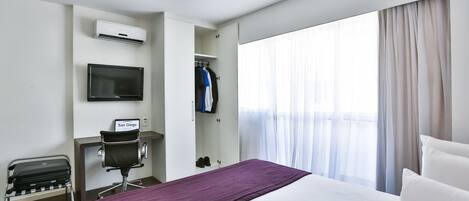A hotel room with a bed, a desk with a chair, a TV, an air conditioning unit, a coat rack, and a window with curtains.
