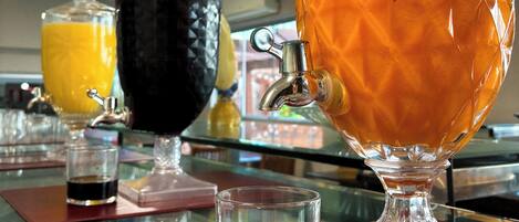 Estación de bebidas con un gran bol de ponche etiquetado como “Laranja com Mamão” y un recipiente más pequeño con bebida negra.