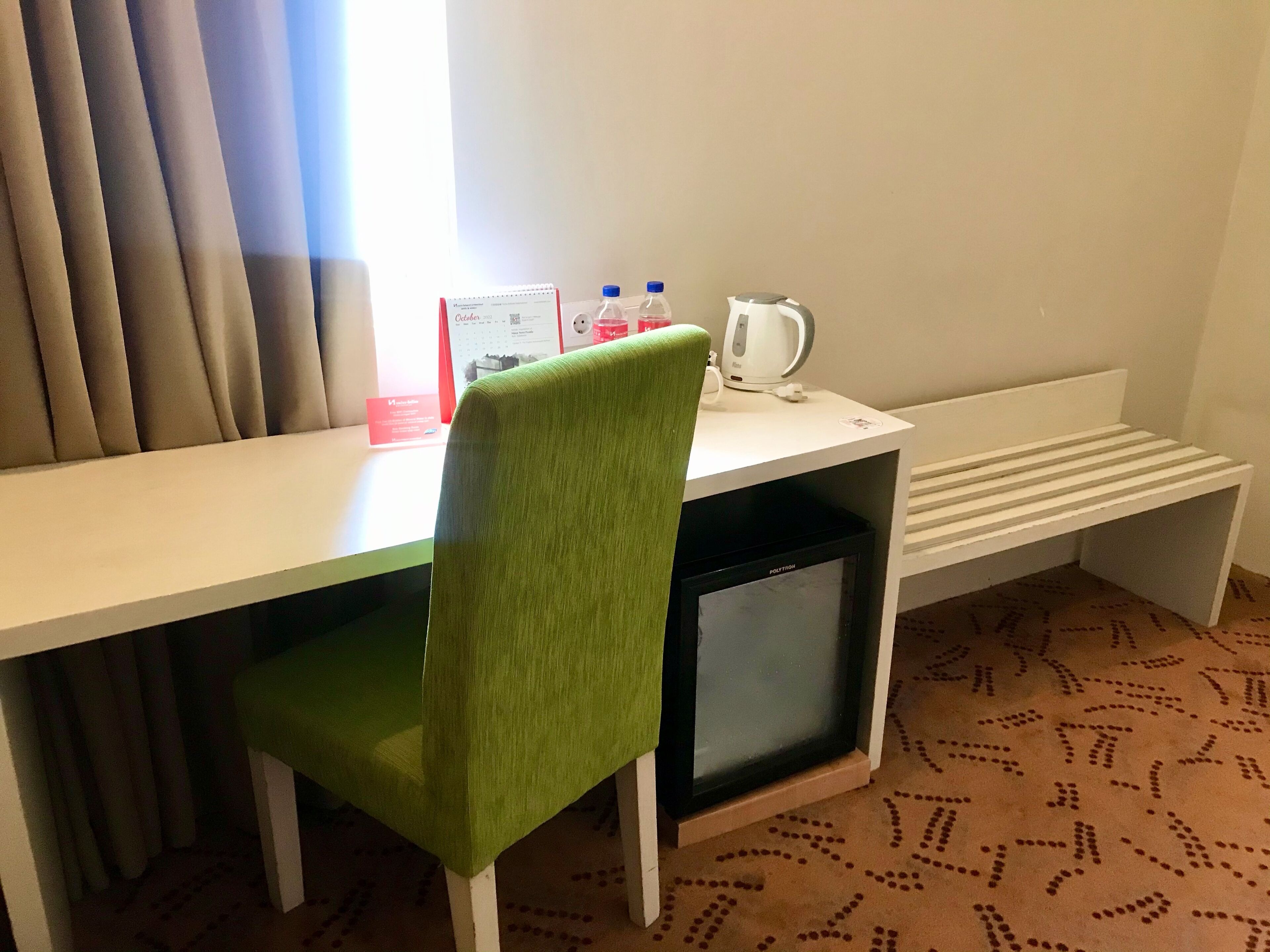 Une chambre d’hôtel équipée d’un bureau, d’une chaise verte, d’une petite télévision, d’une bouilloire et d’une bouteille d’eau.