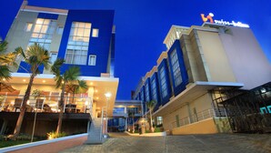 Een modern hotel met een blauw-beige gevel, een bord met 'Swiss-Baillon', en een goed verlicht buitengebied met palmbomen en een overdekt zitgedeelte.