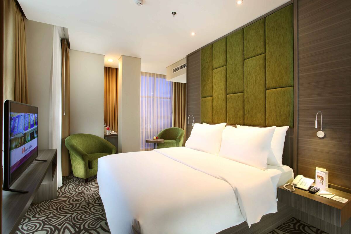 Een hotelkamer met een groot bed, een televisie op een standaard, een groene fauteuil en een tafeltje met een telefoon en een brochure.