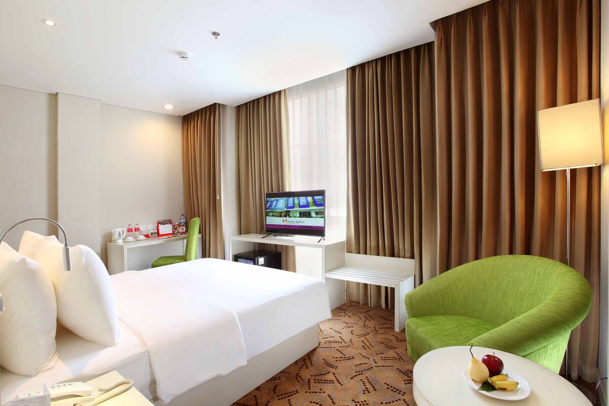 Een hotelkamer met een bed, een bureau met een televisie, een groene fauteuil en een klein tafeltje met fruit.