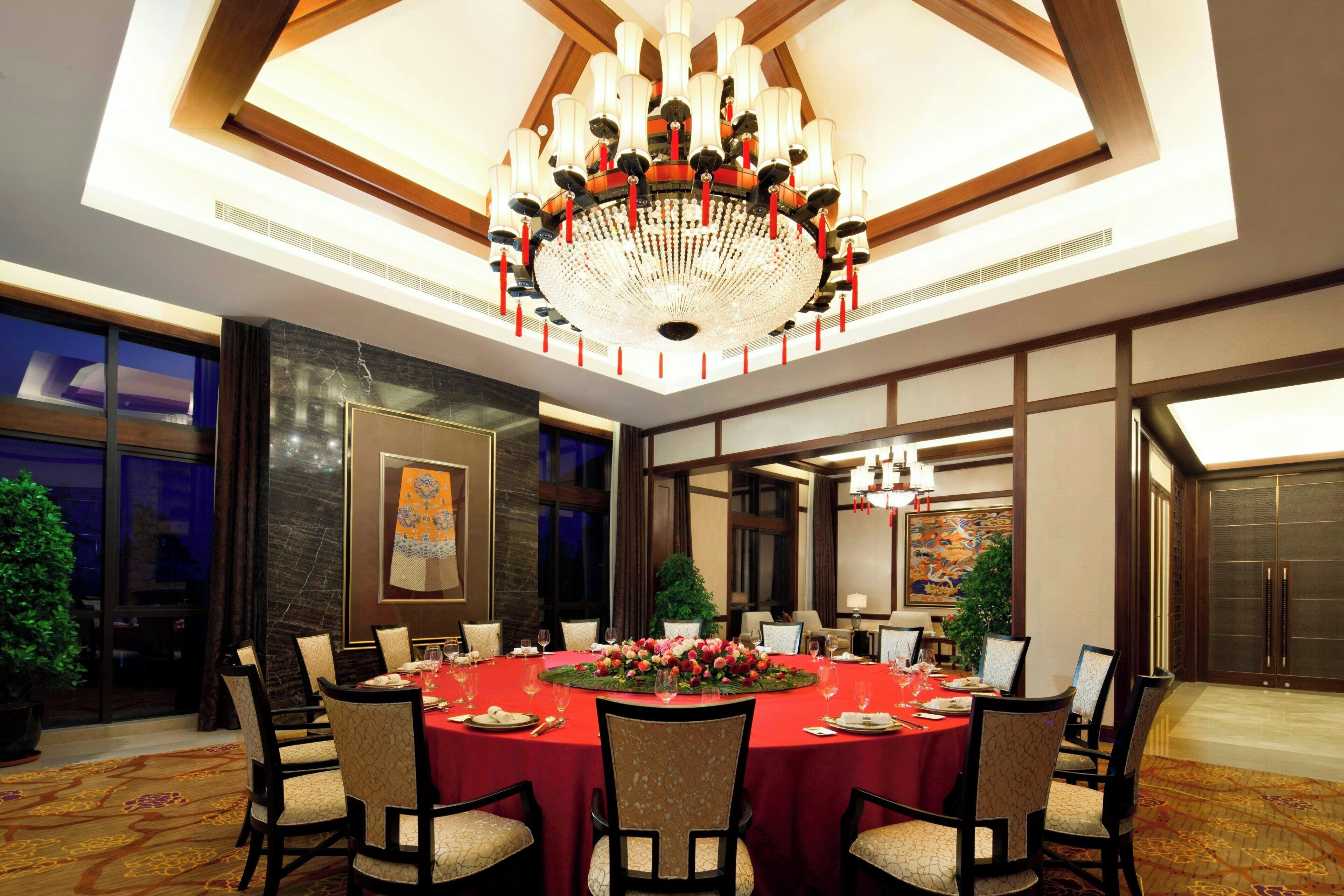 A large, round dining table with a red tablecloth, surrounded by chairs, in a well-lit room with a large chandelier and a view of the outside through large windows.